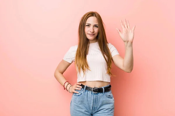 年轻的红发姜女人对一个粉红色的墙微笑欢快显示五号用手指 — 图库照片
