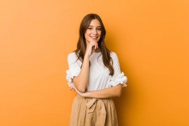 Genç beyaz kadın mutlu ve kendinden emin gülümsüyor, elleriyle çenesine dokunuyor..