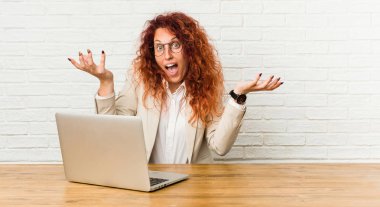 Genç kızıl saçlı kıvırcık kadın bir zafer ya da başarı kutluyor onu dizüstü bilgisayar ile çalışan, o şaşırır ve şok.
