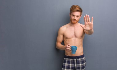 Genç gömleksiz kızıl adam el ele önde. Elinde bir kahve bardağı tutuyor..