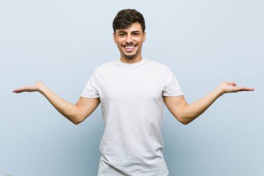 Beyaz bir tshirt giyen genç beyaz adam kolları ile ölçek yapar, mutlu ve kendinden emin hissediyor.