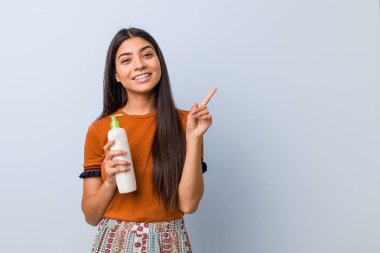 Bir krem şişe tutan genç Arap kadın neşeyle işaret parmağı ile işaret gülümseyerek uzakta.