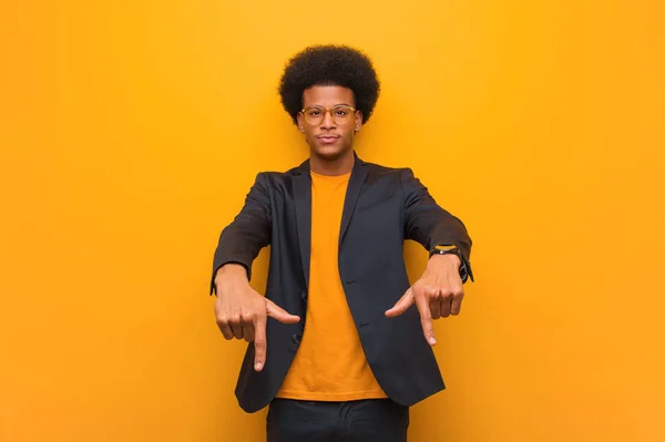 Young business african american man over an orange wall pointing to the bottom with fingers