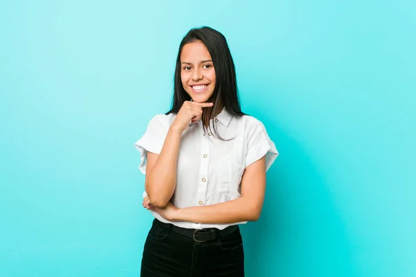 Young hispanic cool woman against a blue wall smiling happy and confident, touching chin with hand.