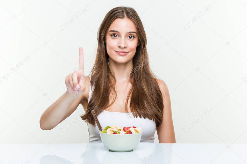 Young caucasian woman eating fruit bowl showing number one with finger.