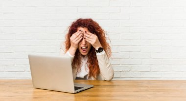 Dizüstü bilgisayarıyla çalışan genç kızıl saçlı kıvırcık kadın elleri ile gözleri kaplıyor, gülümsüyor geniş bir sürpriz bekliyor.