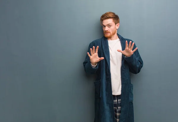 Young redhead man in pajama rejecting something doing a gesture of disgust