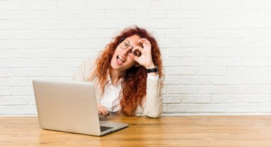 Dizüstü bilgisayarıyla çalışan genç kızıl saçlı kıvırcık kadın göz de ok jest tutmak heyecanlı.