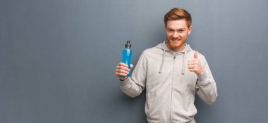 Young redhead fitness man smiling and raising thumb up. He is holding an energy drink.