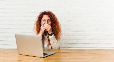 Young redhead curly woman working with her laptop shocked covering mouth with hands.