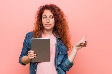 Young caucasian redhead woman holding a tablet smiling cheerfully pointing with forefinger away.