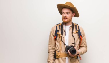 Young redhead explorer man smiling confident and crossing arms, looking up. Holding a camera.