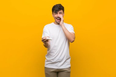 Young hispanic man holding an airplane icon shouting excited to front.