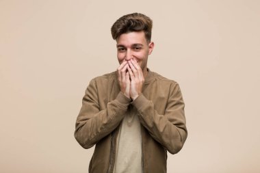 Kahverengi ceketli genç beyaz bir adam bir şeye gülüyor, ağzı ellerle örtüyor..