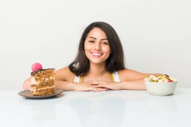 Genç İspanyol kadın pasta ve meyve arasında seçim yapıyor. Mutlu, gülümseyen ve neşeli..