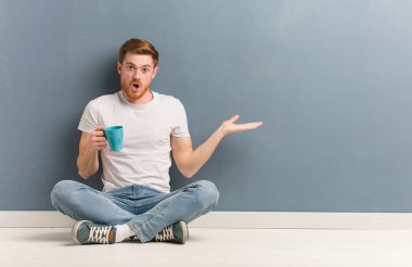 Genç, kızıl saçlı bir öğrenci, elinde bir şey tutarak yerde oturuyor. Elinde bir kahve fincanı var..