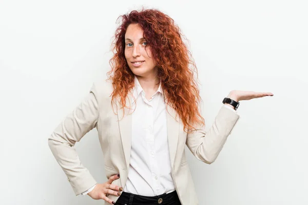 Young natural redhead business woman isolated against white background showing a copy space on a palm and holding another hand on waist.