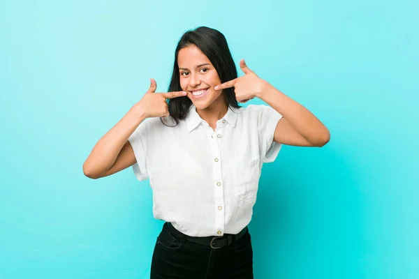 Young hispanic cool woman against a blue wall smiles, pointing fingers at mouth.