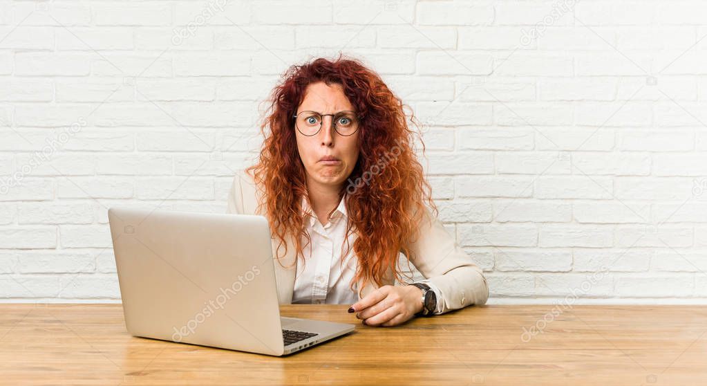 Young redhead curly woman working with her laptop shrugs shoulders and open eyes confused.