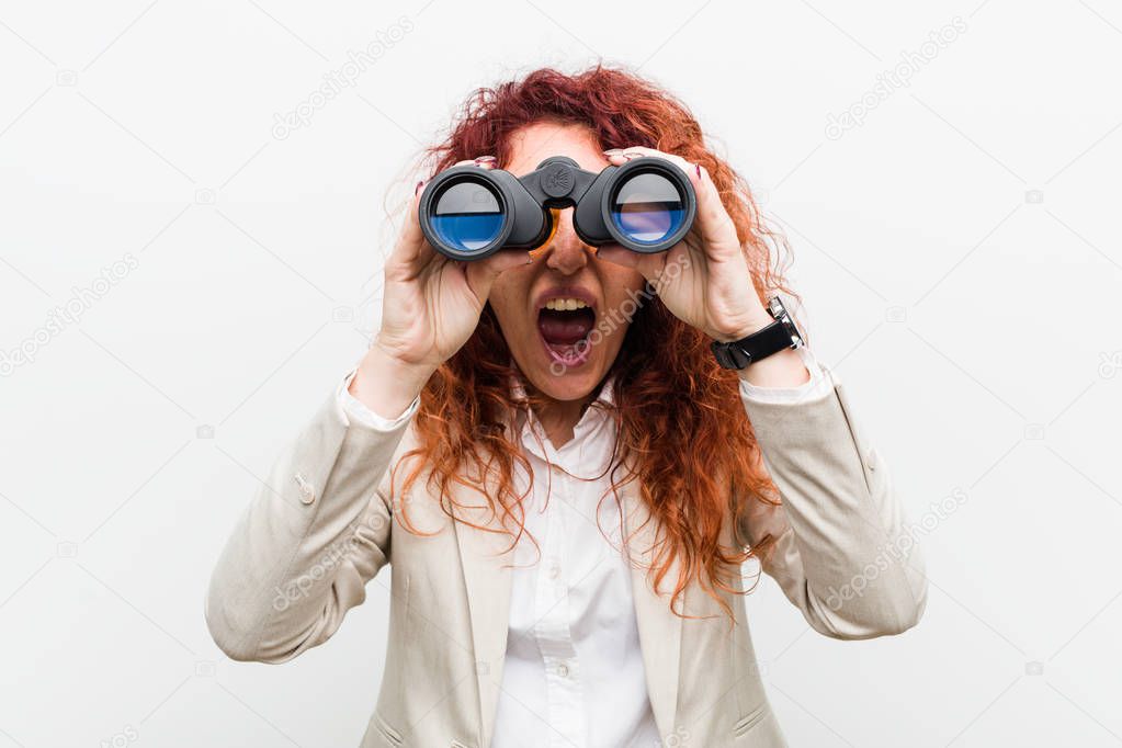 Young caucasian business redhead woman holding a binoculars shouting excited to front.