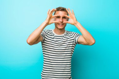 Mavi duvara yaslanmış genç beyaz bir adam bir başarı fırsatı bulmak için gözlerini açık tutuyor..