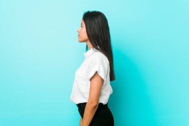 Genç Latin asıllı havalı kadın mavi bir duvara yaslanmış, sola bakıyor, yandan poz veriyor.