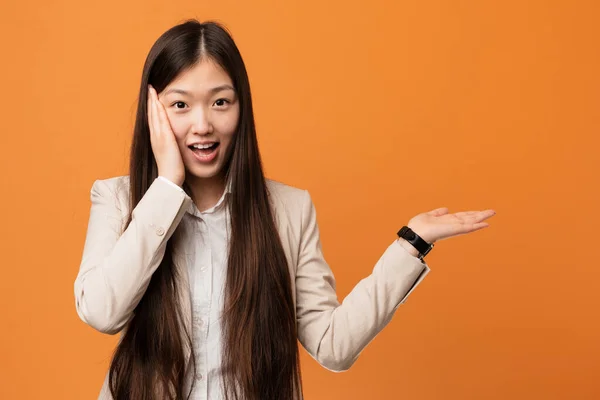 Young business chinese woman holds copy space on a palm, keep hand over cheek. Amazed and delighted.