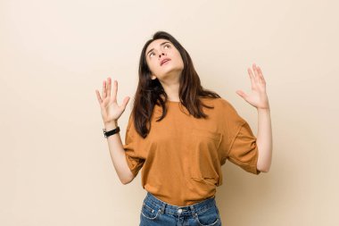 Genç esmer kadın bej bir arka planda gökyüzüne haykırıyor, yukarı bakıyor, hayal kırıklığına uğramış..