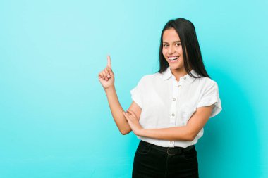 Mavi bir duvara yaslanmış, işaret parmağıyla mutlu bir şekilde gülümseyen genç İspanyol havalı kadın..