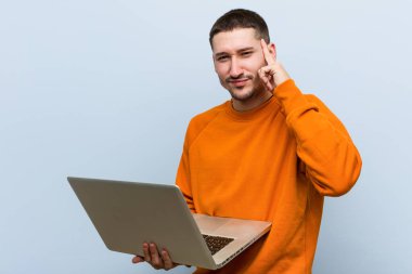 Genç, beyaz bir adam elinde dizüstü bilgisayarla şakağını işaret ediyor. Düşünüyor, bir göreve odaklanıyor..