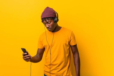Bir telefon ile müzik dinlerken bir şapka giyen sarı bir arka plan karşı duran genç Afrikalı adam