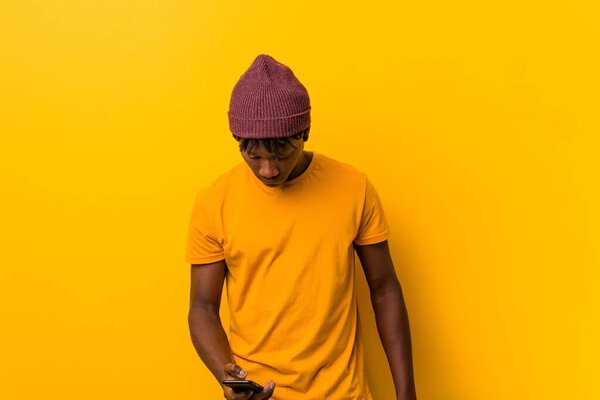 Young african man standing against a yellow background wearing a hat and using a phone
