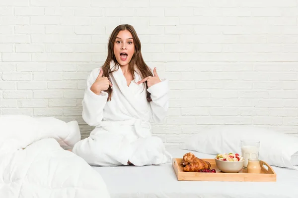 Young caucasian woman on the bed surprised pointing with finger, smiling broadly.