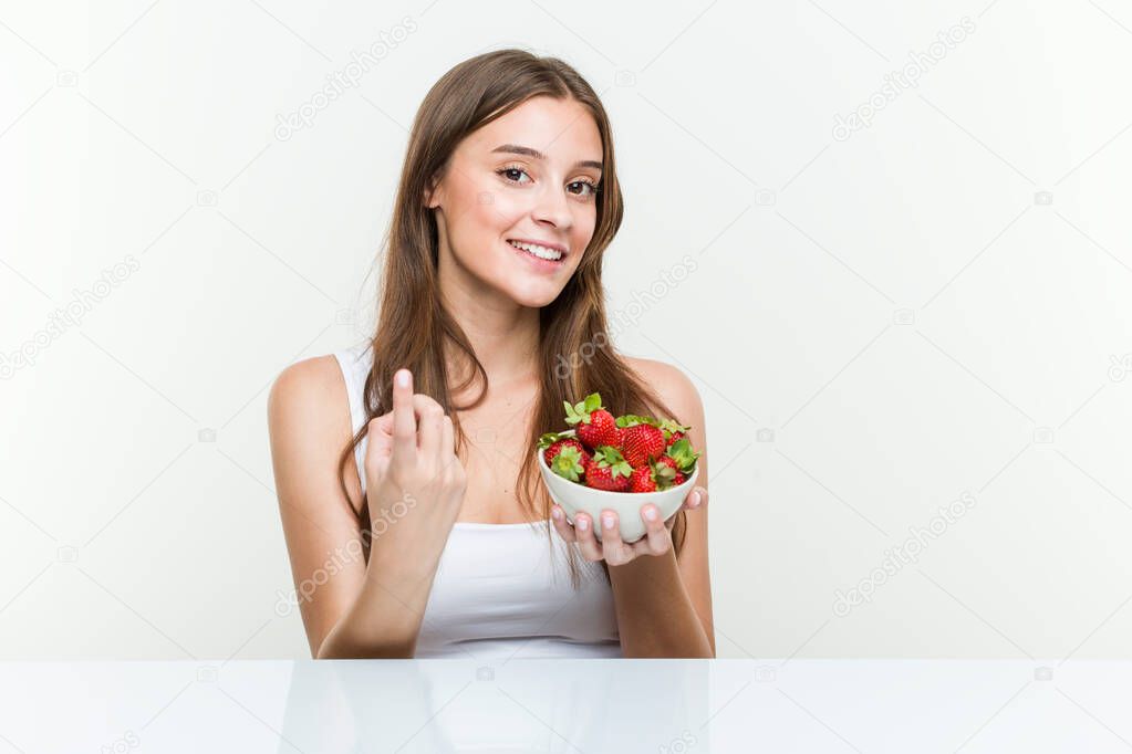 Young caucasian woman holding a strawberries bowl pointing with finger at you as if inviting come closer.