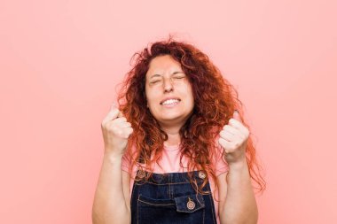 Genç, kızıl saçlı, kot pantolon giyen güzel bir kadın yumruğunu kaldırıyor, mutlu ve başarılı hissediyor. Zafer konsepti.