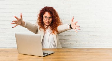 Genç kızıl saçlı, laptopuyla çalışan kıvırcık kadın kameraya sarıldığı için kendinden emin..