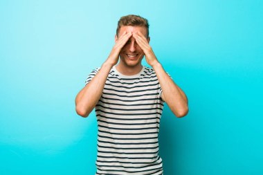 Mavi bir duvarın karşısında genç beyaz bir adam elleriyle gözlerini kapatıyor, gülümseyerek bir sürpriz bekliyor..