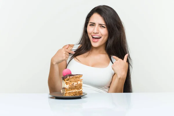 Young hispanic woman eating a cake surprised pointing at himself, smiling broadly.