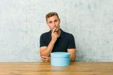 Masada hediye kutusu tutan beyaz bir adam rahatlamış fotokopi odasına bakan bir şey düşünüyor..