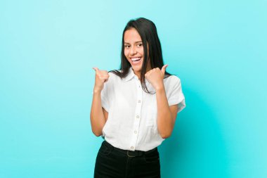 Genç, İspanyol asıllı, havalı bir kadın, mavi bir duvara yaslanmış, başparmağını kaldırıyor, gülümsüyor ve kendinden emin..