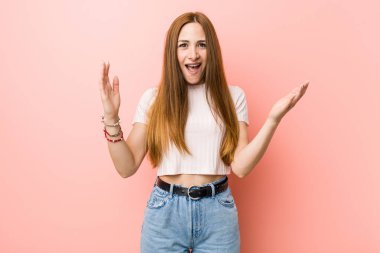 Genç kızıl saçlı kadın pembe bir duvara yaslanmış. Hoş bir sürpriz yapıyor. Heyecanlı ve el kaldırıyor..