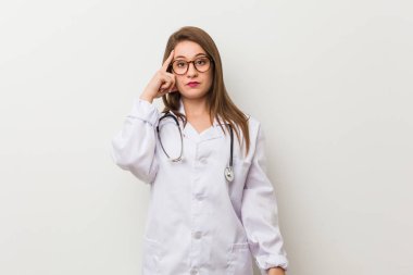 Genç doktor kadın, beyaz bir duvara yaslanmış, parmağıyla şakağı işaret ediyor, düşünüyor ve bir göreve odaklanıyor..