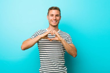 Mavi bir duvarın karşısında gülümseyen ve elleriyle kalp şeklini gösteren genç beyaz bir adam..