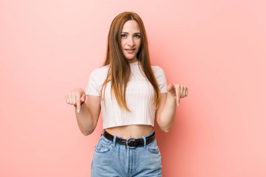 Kızıl saçlı genç kadın pembe bir duvara karşı parmaklarıyla işaret ediyor, olumlu bir his..
