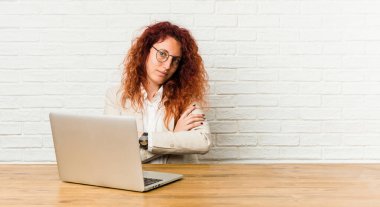 Dizüstü bilgisayarıyla çalışan genç, kızıl saçlı kıvırcık kadın. Kendine güveniyor, kararlılıkla kollarını kavuşturuyor..