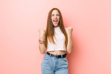 Genç kızıl saçlı kadın pembe bir duvara yaslanmış kaygısız ve heyecanlı tezahüratlar yapıyor. Zafer konsepti.