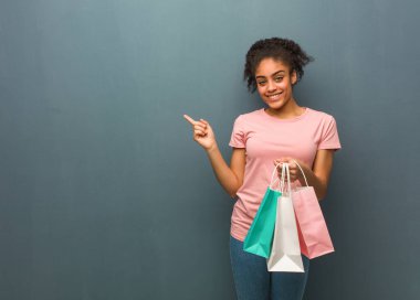 Genç siyah kadın gülümsüyor ve yan tarafı işaret ediyor. Elinde bir alışveriş çantası var..