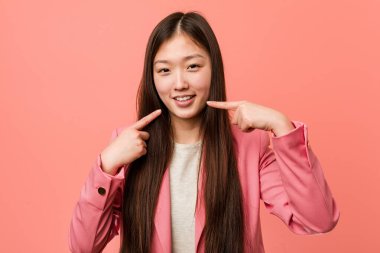 Pembe takım elbiseli genç Çinli kadın gülümsüyor, parmaklarını ağzına doğrultuyor..