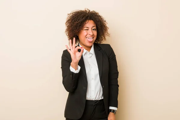 Young business african american woman winks an eye and holds an okay gesture with hand.