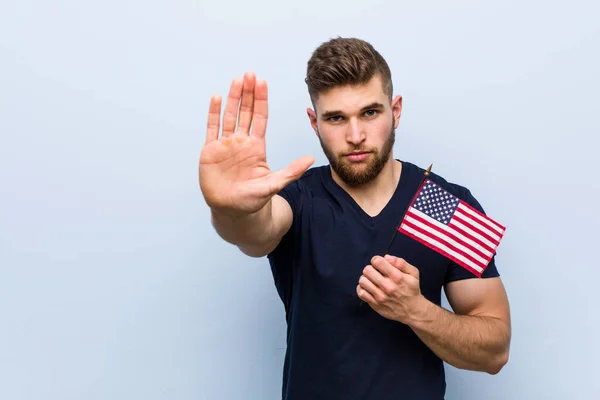 Young caucasian man holding a united states flag standing with outstretched hand showing stop sign, preventing you.